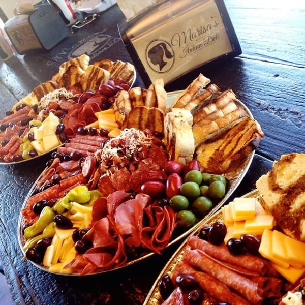 Three large platters of Italian deli meats, cheeses, olives, and bread on a dark wooden table.