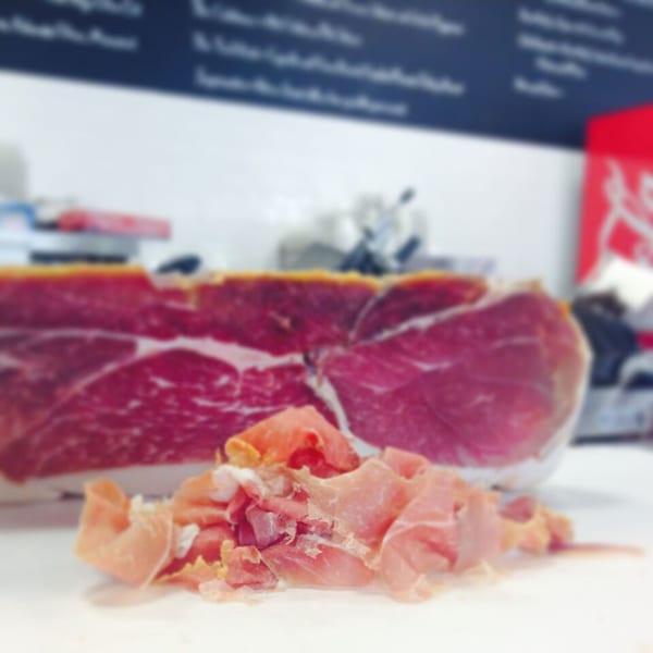 A large cut of prosciutto with a pile of thinly sliced deli meat in the foreground.