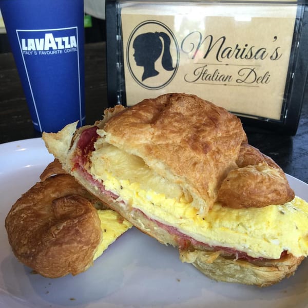 A croissant breakfast sandwich with egg, ham, and cheese, with a Lavazza coffee cup and Marisa's Italian Deli sign in the background.